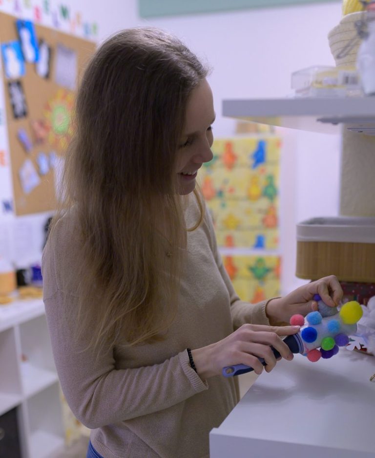 Frau mit Pompons Eine Person ordnet farbige Spielzeuge auf einem Regal in einem Raum.