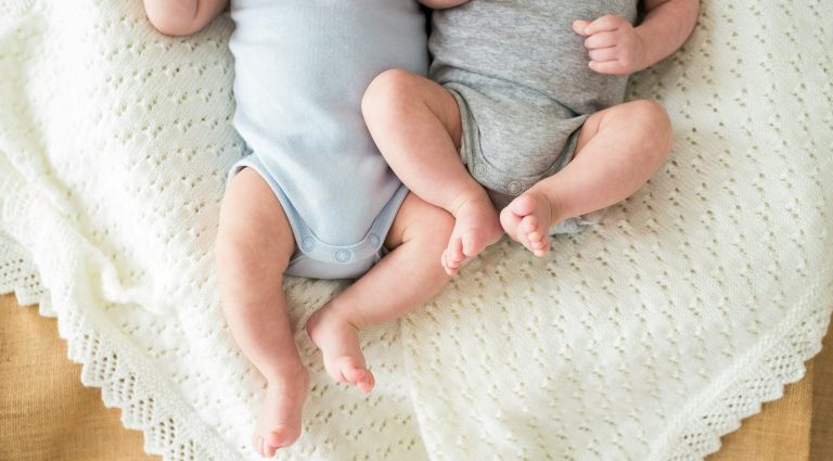 Zwei Babys in bodenlangen Stramplern liegen auf einer weichen Decke.