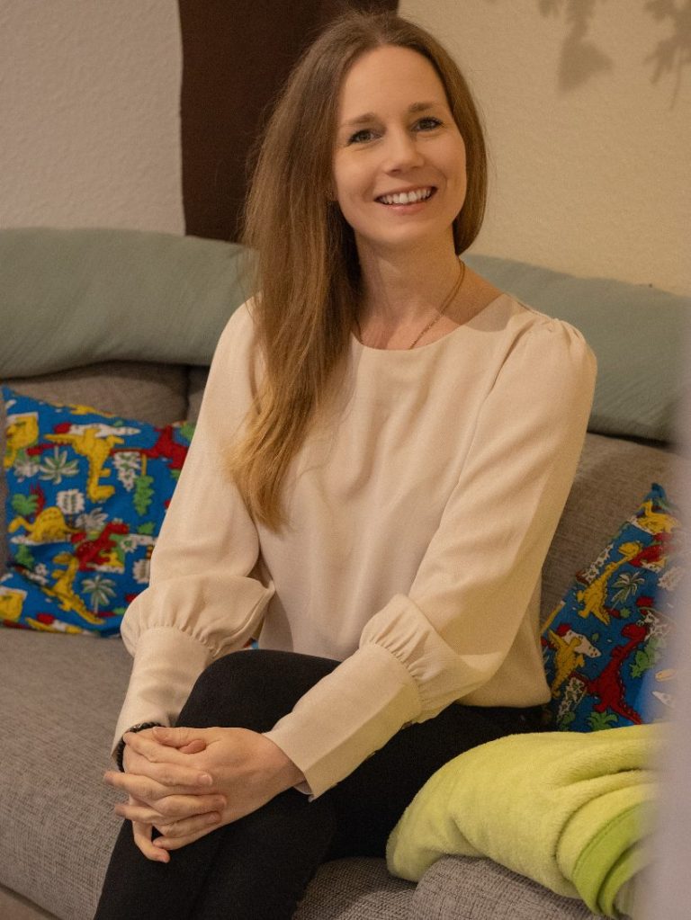 Lächelnde Frau mit langen Haaren sitzt auf einem Sofa zwischen bunten Kissen.