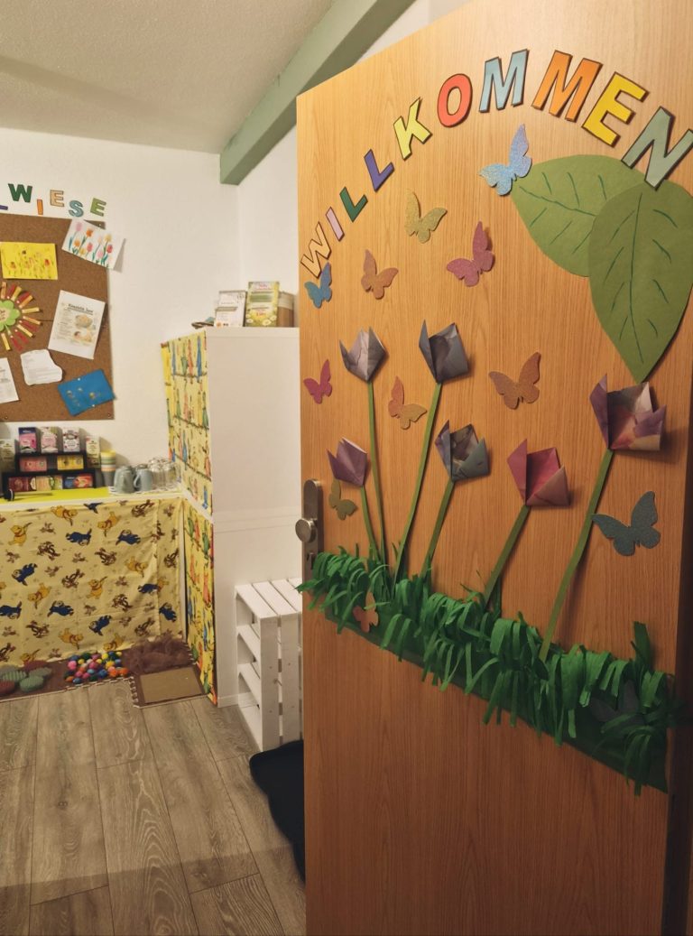 Holztür mit "Willkommen"-Schriftzug, umgeben von Blumen und Schmetterlingen.