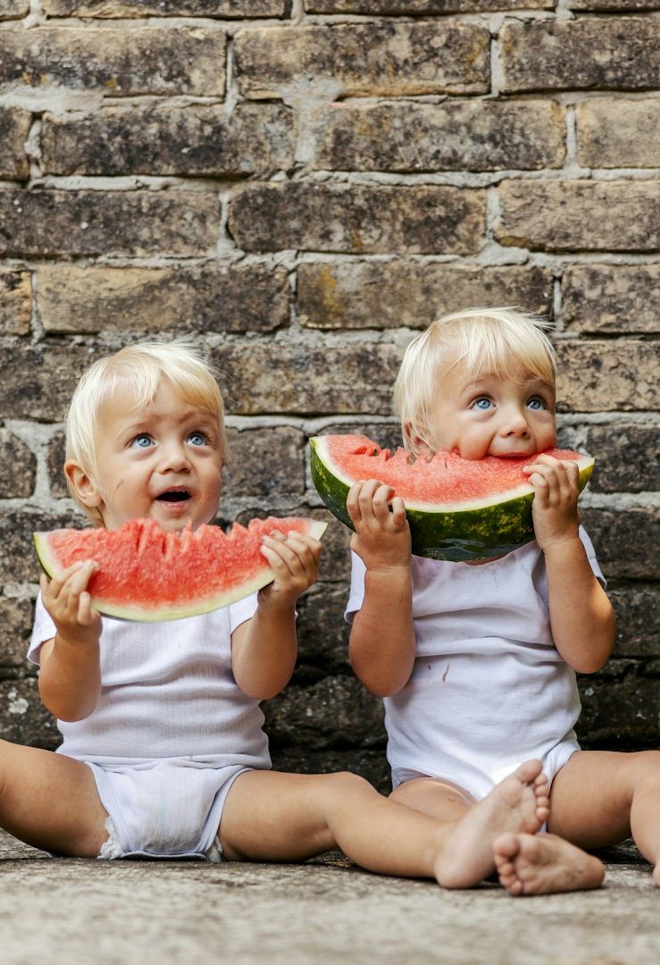 Zwei kleine Kinder mit blonden Haaren sitzen und essen Wassermelone.