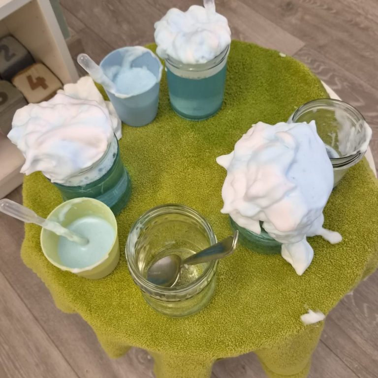 Experimentieren für Kinder Fünf Gläser mit bunten Flüssigkeiten und Schaum auf einem grünen Tisch.