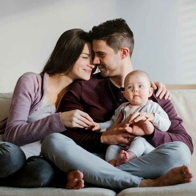 Familienberatung Lächelnde Familie mit einem Baby, auf einer Couch sitzend und sich umarmend.