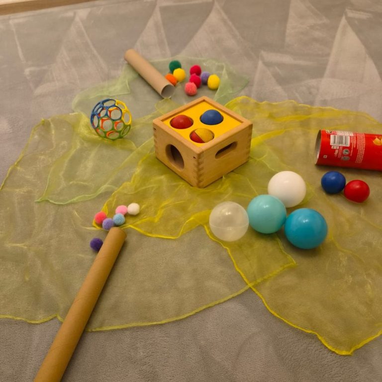 Montessori Pädagogik Spielaufbau für die kleinen Krabbler Bunte Spielzeuge und Materialien liegen auf einer gelben Unterlage verteilt.