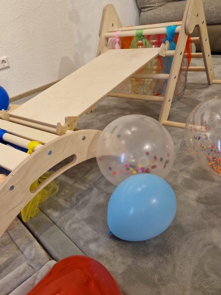 Montessori Kletterset Woodscraft bei der Krabbelwiese Baden-Baden Holzspielzeug mit einer Rutsche, Luftballons und bunten Kugeln auf dem Boden.